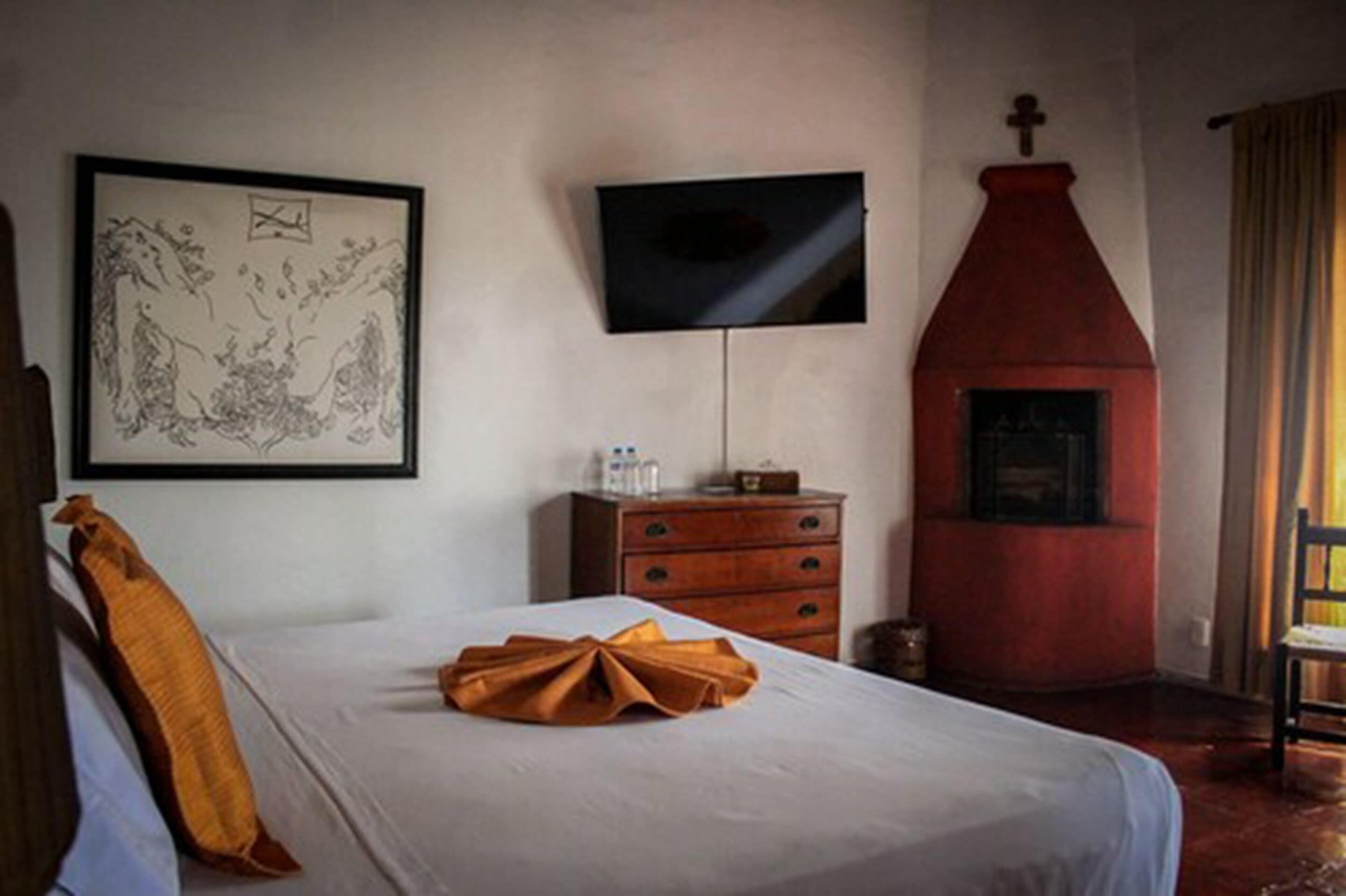 A hotel room with a bed, a flat-screen TV, a wooden dresser, a red fireplace, and a framed artwork on the wall.