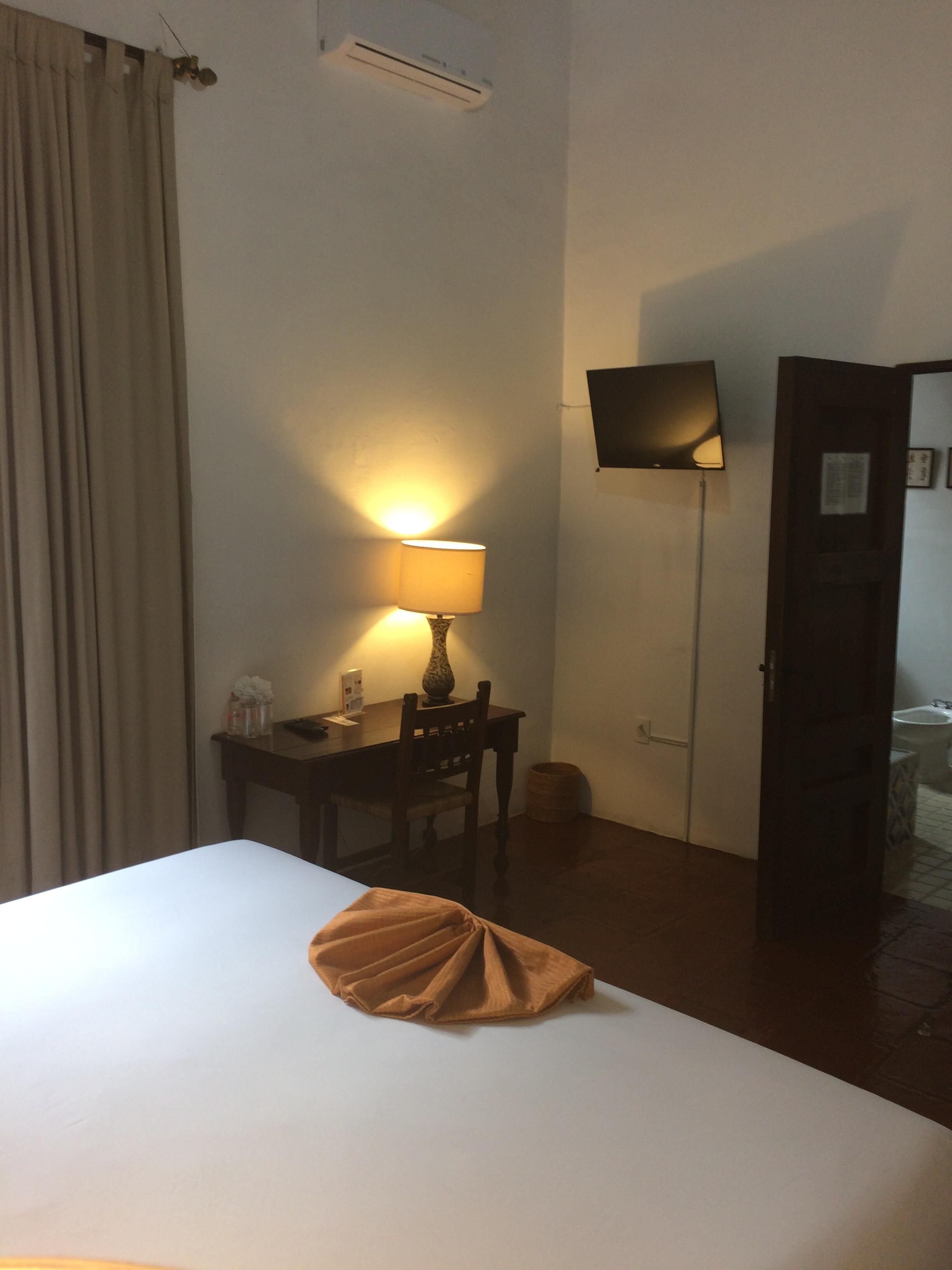 A hotel room with a bed, a desk with a lamp, a television on the wall, and a door leading to a bathroom.