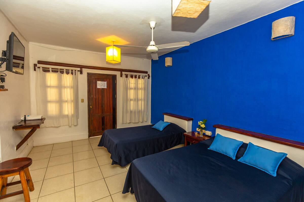 A room with two beds, a ceiling fan, a small table with a lamp, and a window with curtains.