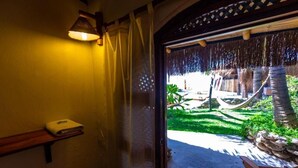 Une porte mène à un hamac et à une structure dotée d’un toit de chaume. Une étagère y est fixée, sur laquelle se trouve un objet blanc, et une lampe est accrochée au mur.