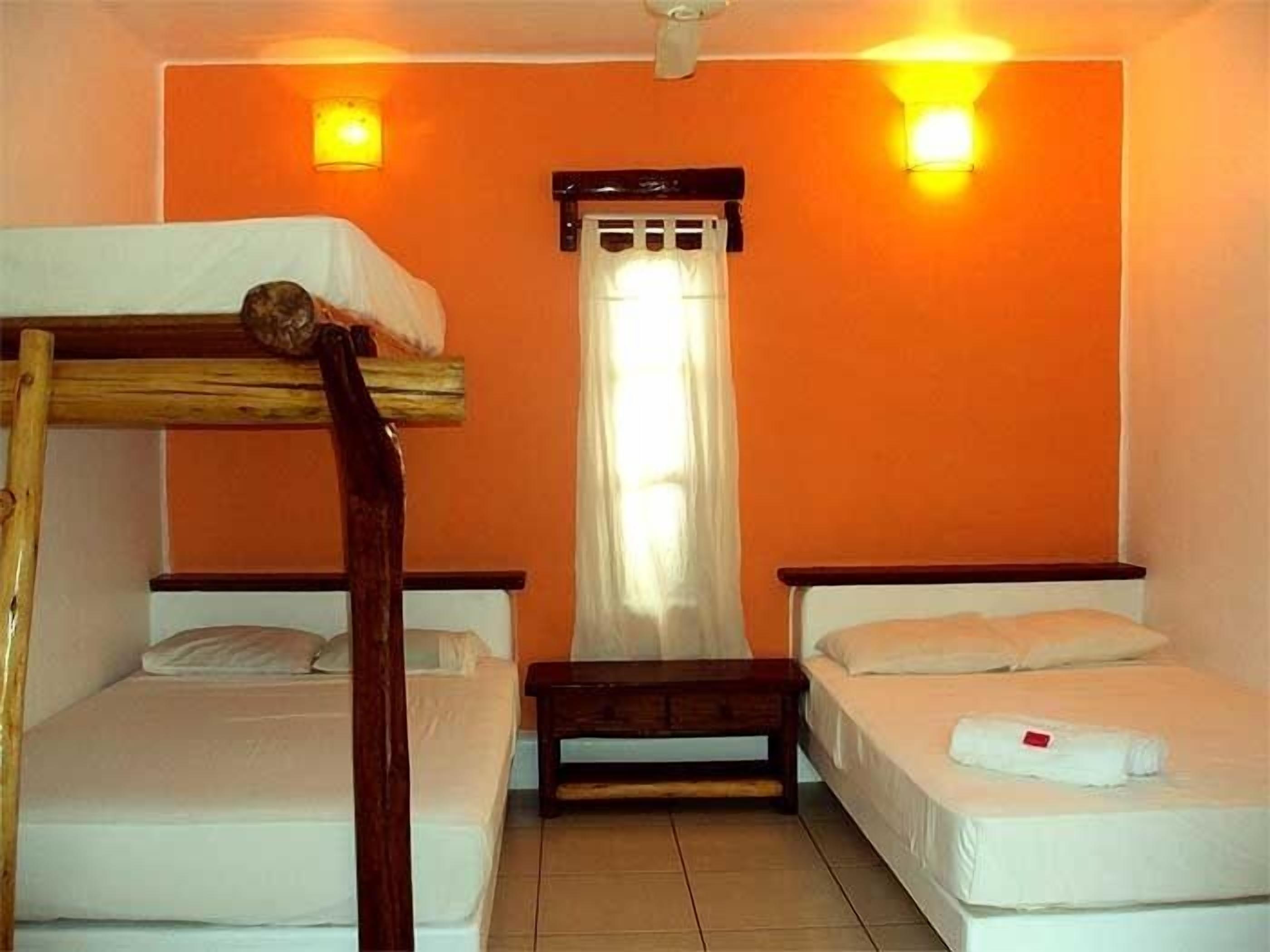A room with two beds, one with a wooden headboard and the other with a metal frame. There is a bench between the beds, and a window with curtains.