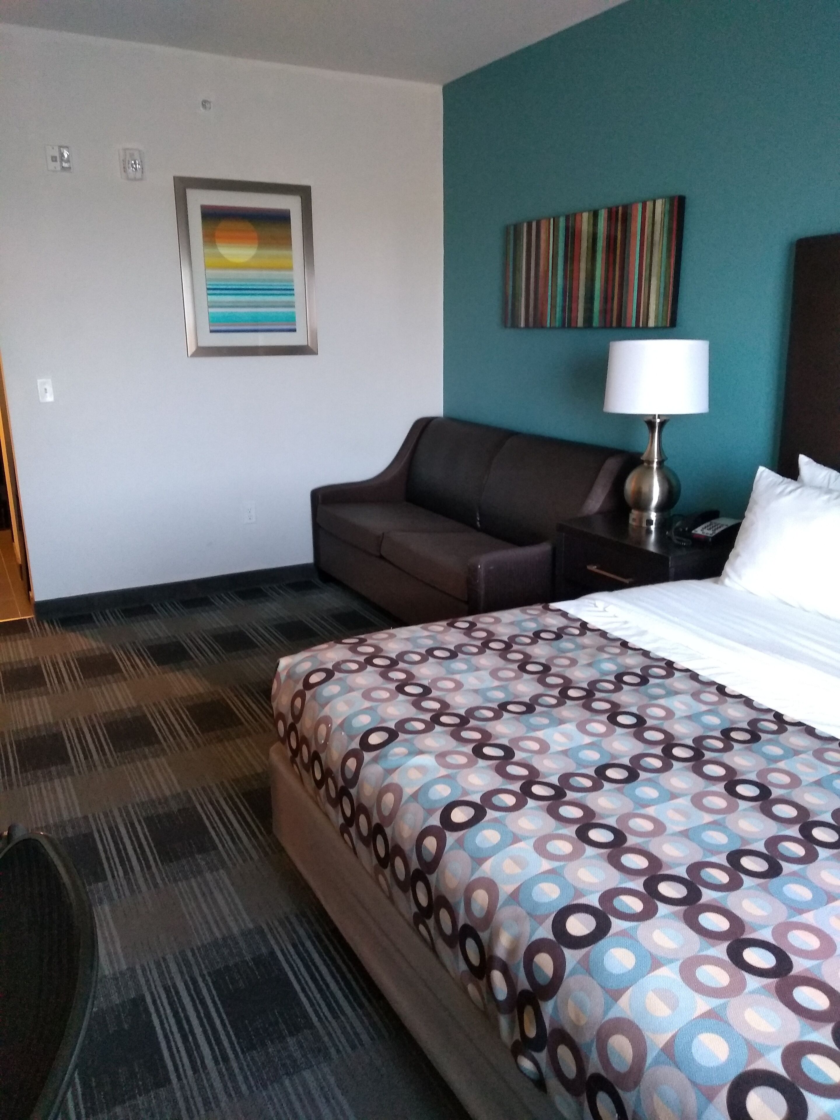 A hotel room with a bed, a sofa, a nightstand with a lamp, a painting on the wall, and a carpeted floor.