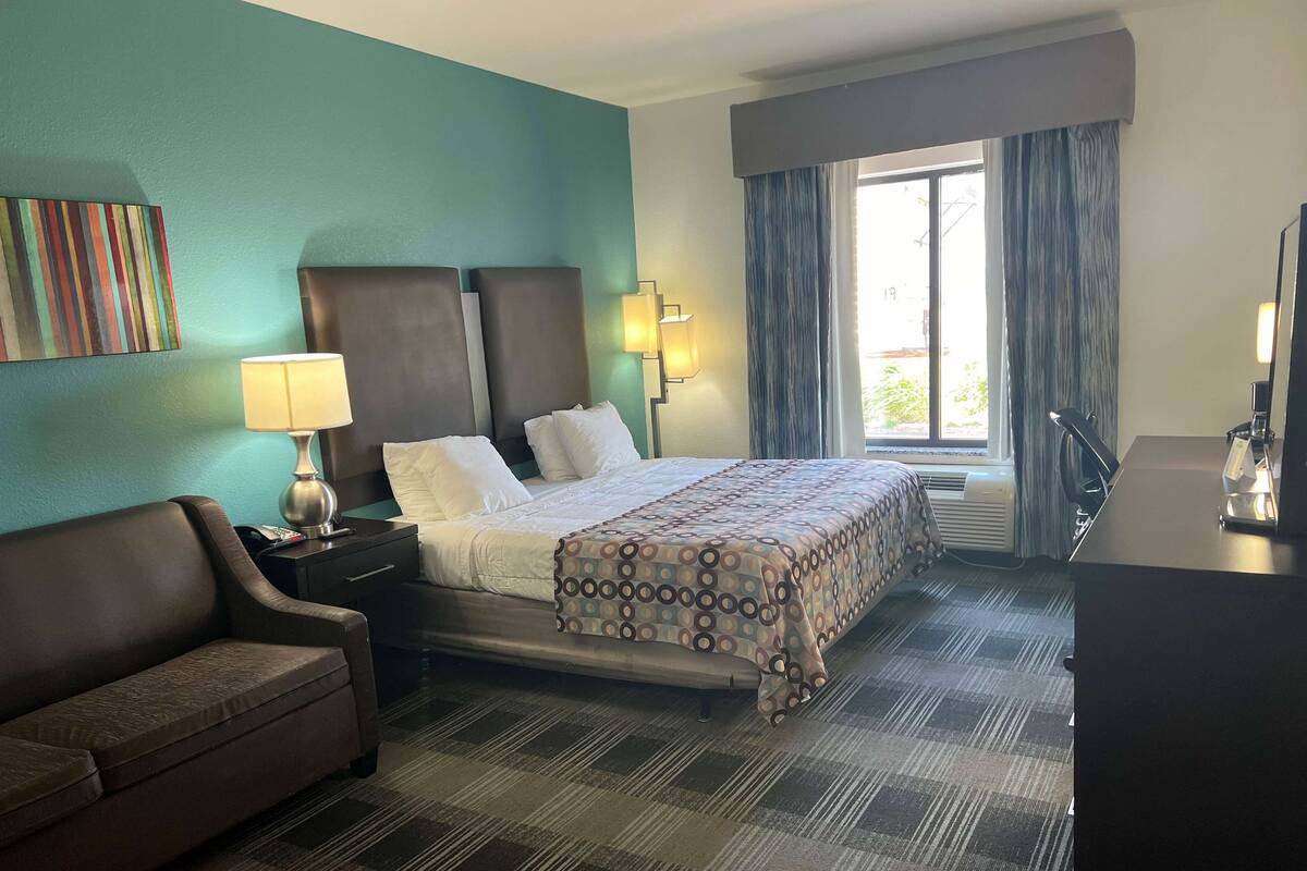 A hotel room with a bed, a nightstand, a desk, a chair, a window with curtains, and a wall with a colorful artwork.