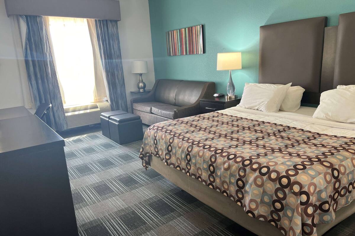 A hotel room with a large bed, a desk, a chair, a nightstand, a lamp, and a window with curtains.