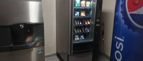 Una máquina expendedora con aperitivos y el logotipo de Pepsi, un refrigerador de acero inoxidable y un piso de baldosas.