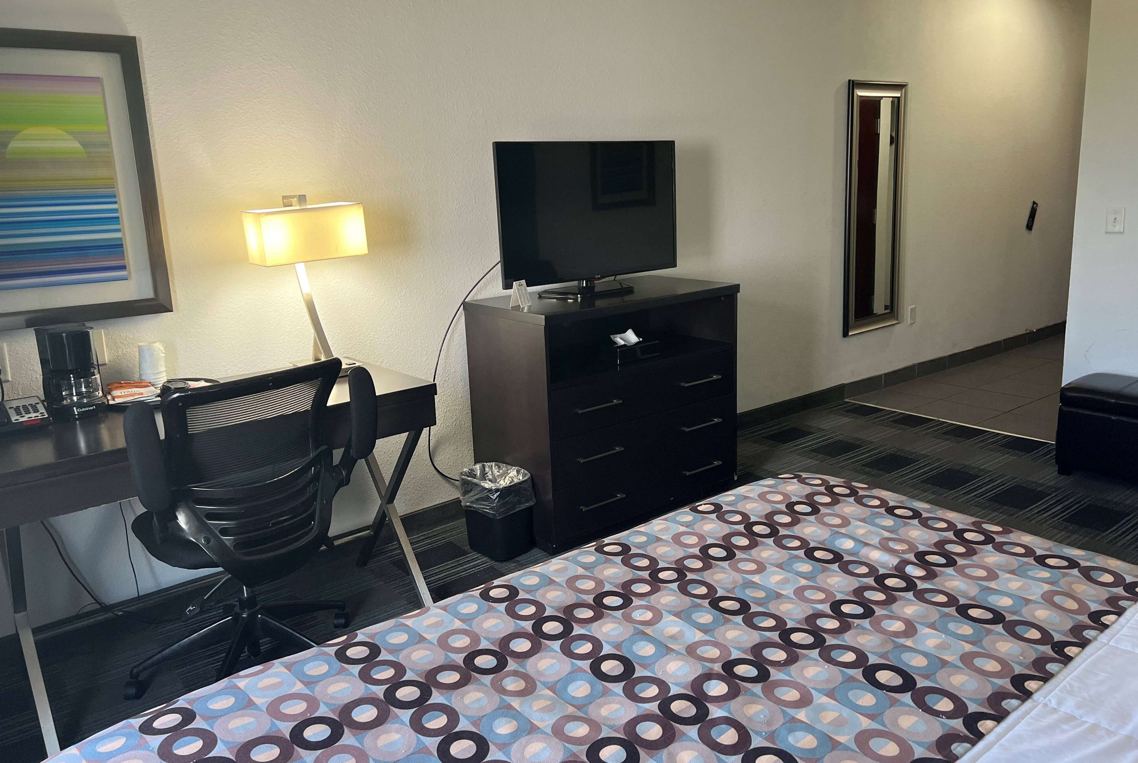 A hotel room with a bed, a desk with a chair, a television, a dresser, a trash bin, and a mirror.