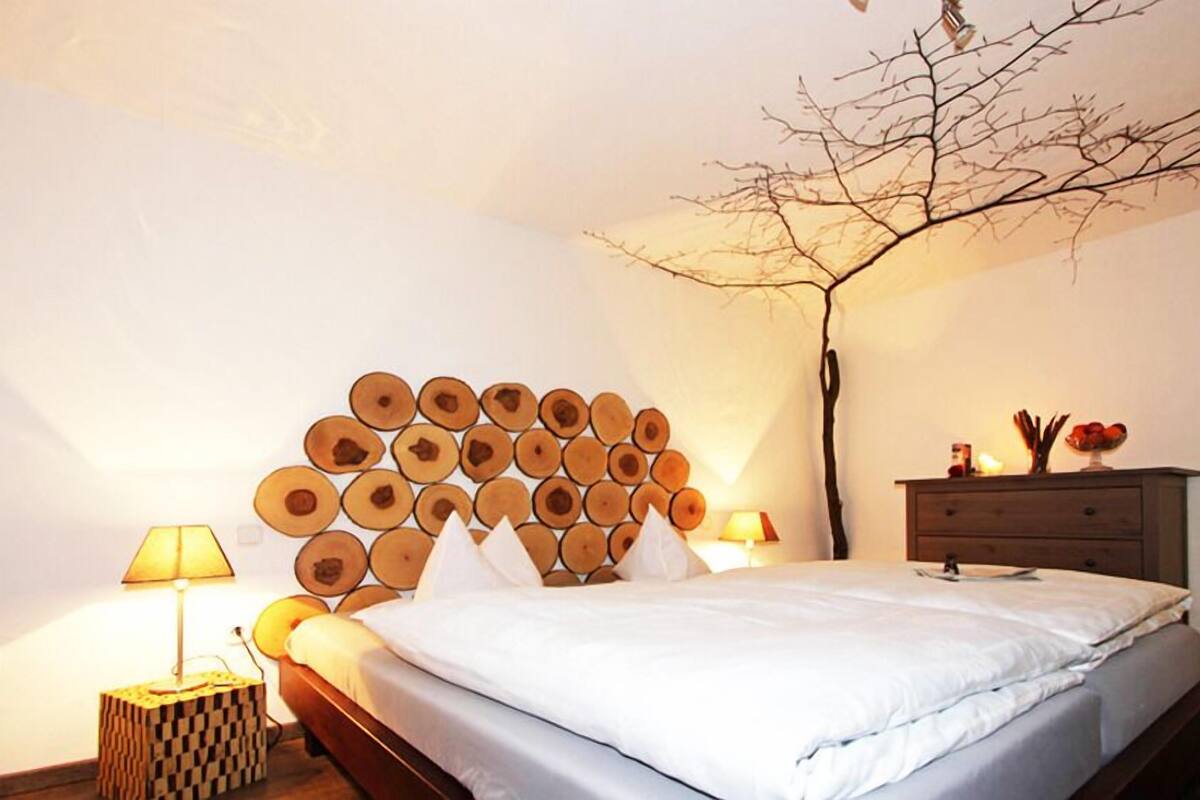 Un lit bien fait, avec une tête de lit faite de rondelles de bois disposées en cercle, une lampe de chevet, une petite table d’appoint en bois et une commode agrémentée d’objets décoratifs.