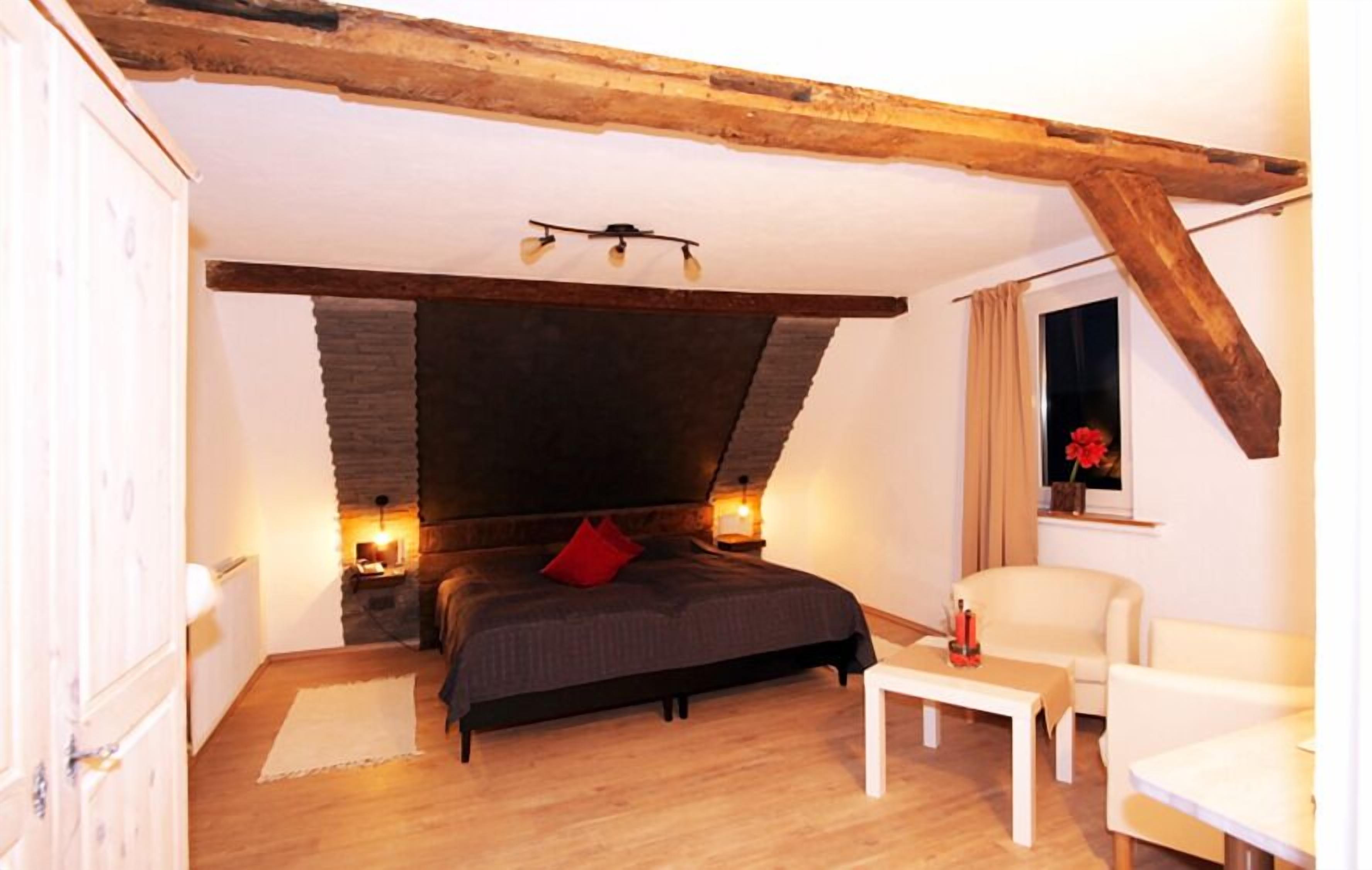 Un dormitorio con chimenea de piedra, una cama con almohada roja, una silla blanca y una mesita pequeña.