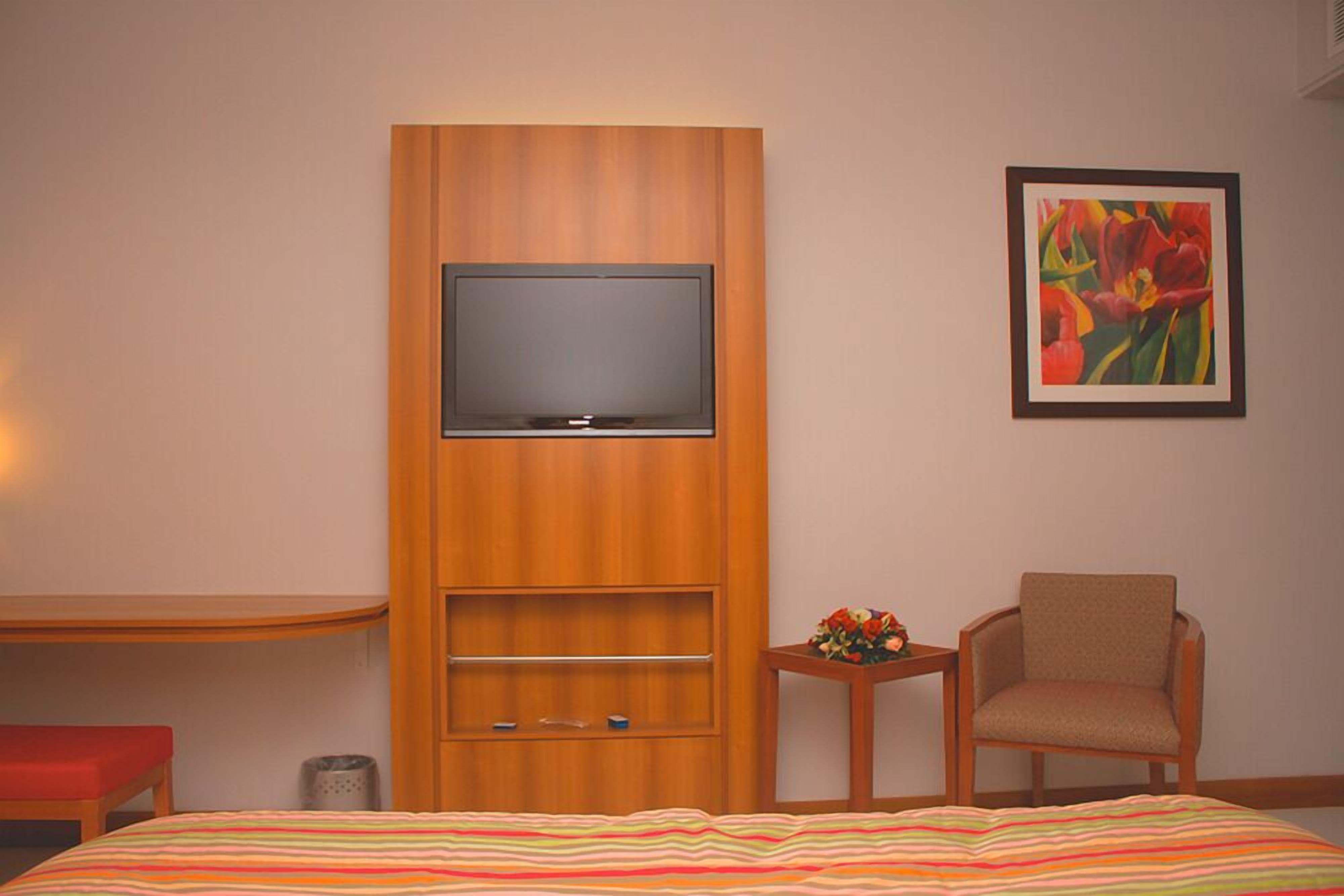 Ein Hotelzimmer mit einem Bett, einem an einer Holzkommode befestigten Fernseher, einem Sessel, einem kleinen Tisch mit Blumenarrangement und einem Bild an der Wand.