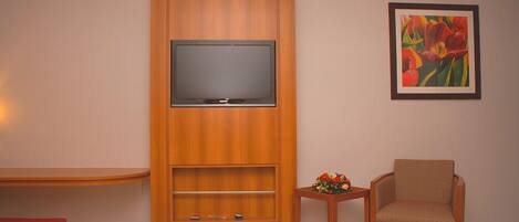 A hotel room with a bed, a TV mounted on a wooden cabinet, a chair, a small table with a flower arrangement, and a painting on the wall.