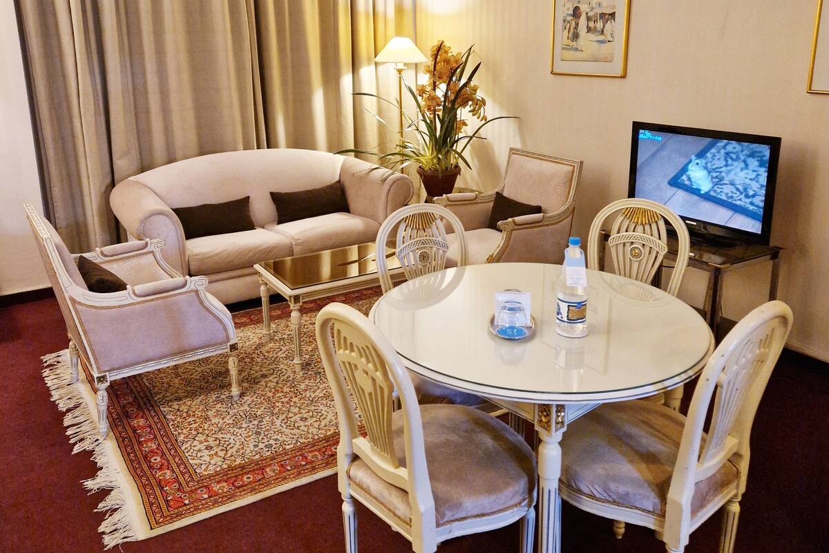 Een hotelkamer met een ronde eettafel, witte stoelen, een bank, een televisie en een potplant.