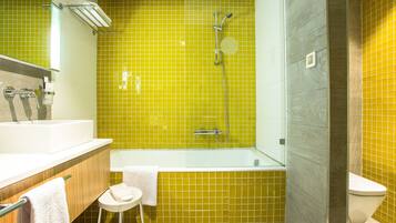 Un baño con pared de azulejos amarillos, una bañera blanca, un inodoro blanco y un taburete pequeño con una toalla.