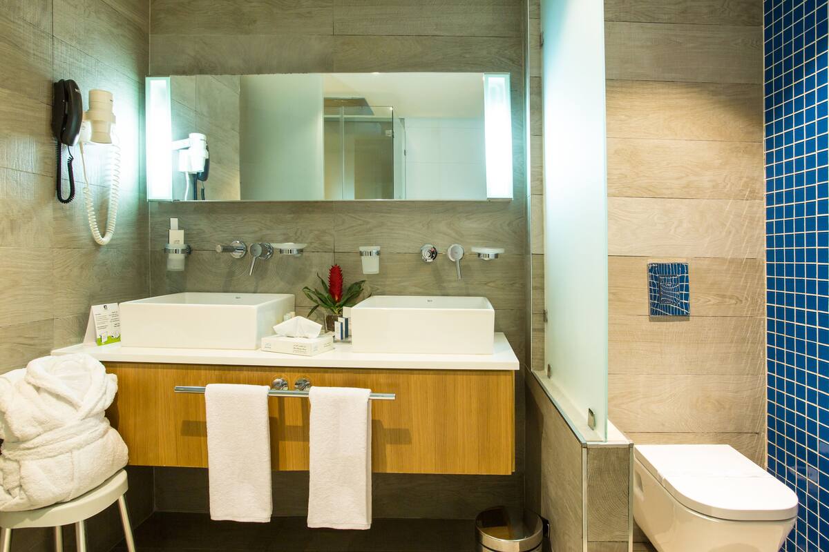 Een moderne badkamer met een houten wastafelmeubel, twee wastafels, een spiegel, een telefoon en een toilet.