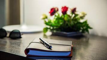 Un cuaderno con un bolígrafo sobre una superficie de madera, un par de gafas y una planta en maceta con flores rojas al fondo.