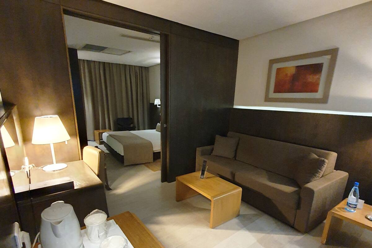 Hotelkamer met een bed, een bank, een salontafel, een bijzettafeltje met een fles en een dienblad met een waterkoker en glazen.