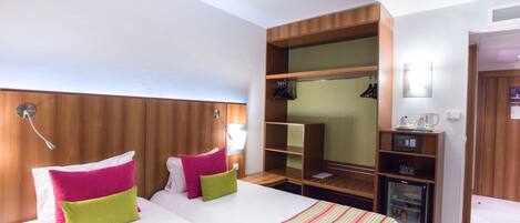 A hotel room with a large bed, two pillows, a bedside table, a wardrobe, and a small refrigerator.