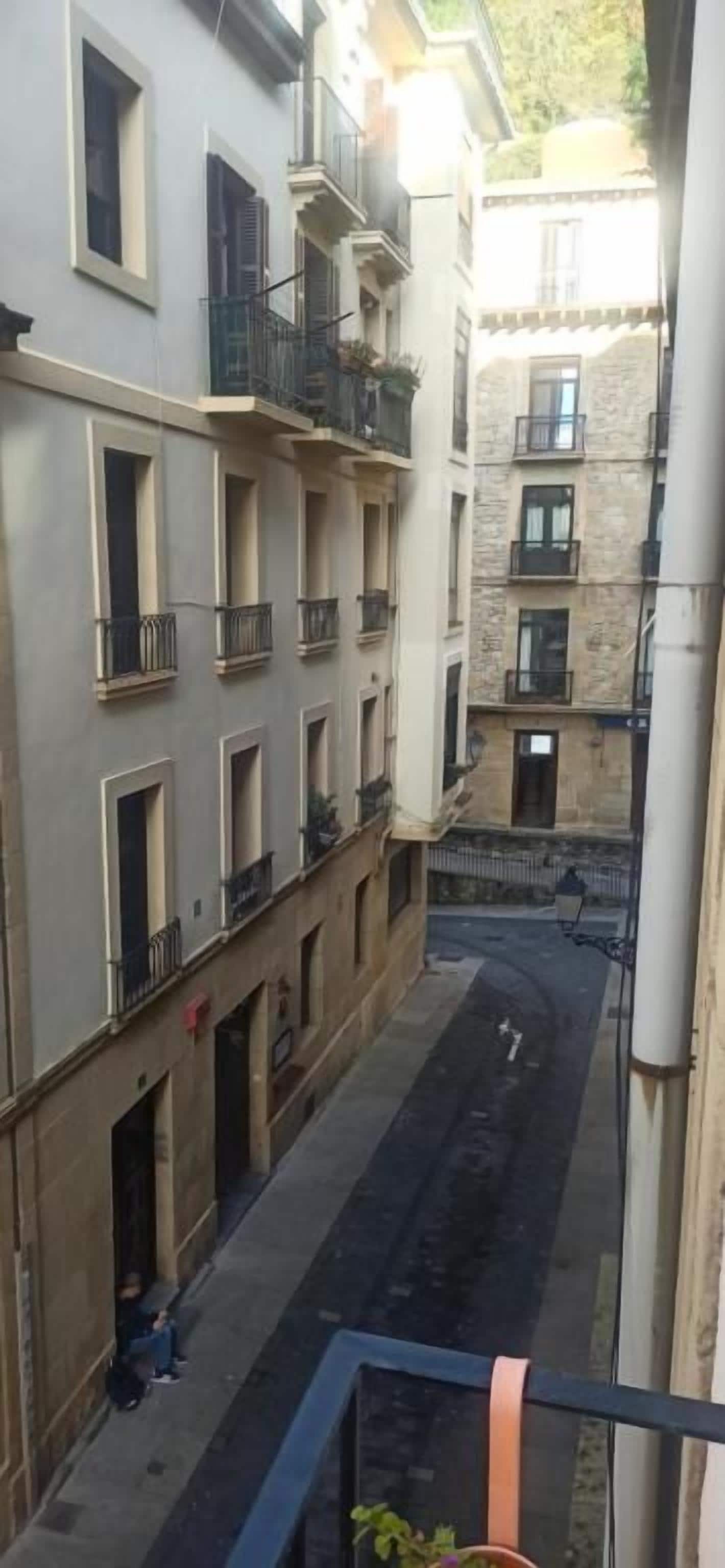 Una calle estrecha entre edificios con balcones, cielo despejado y un peatón.