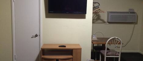 A room with a closed door, a television mounted on the wall, a small wooden cabinet, a chair, and a trash bin.