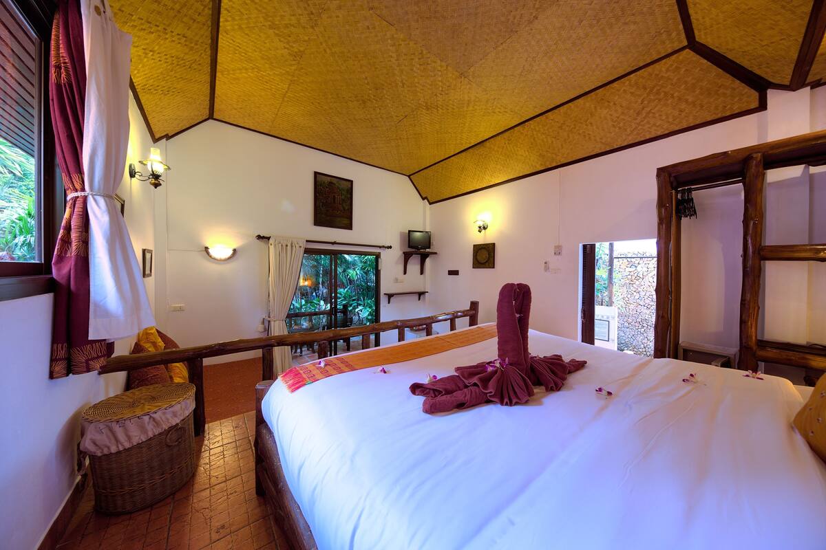 A hotel room with a bed, a television, a window with curtains, a wooden bench, and a wicker basket.