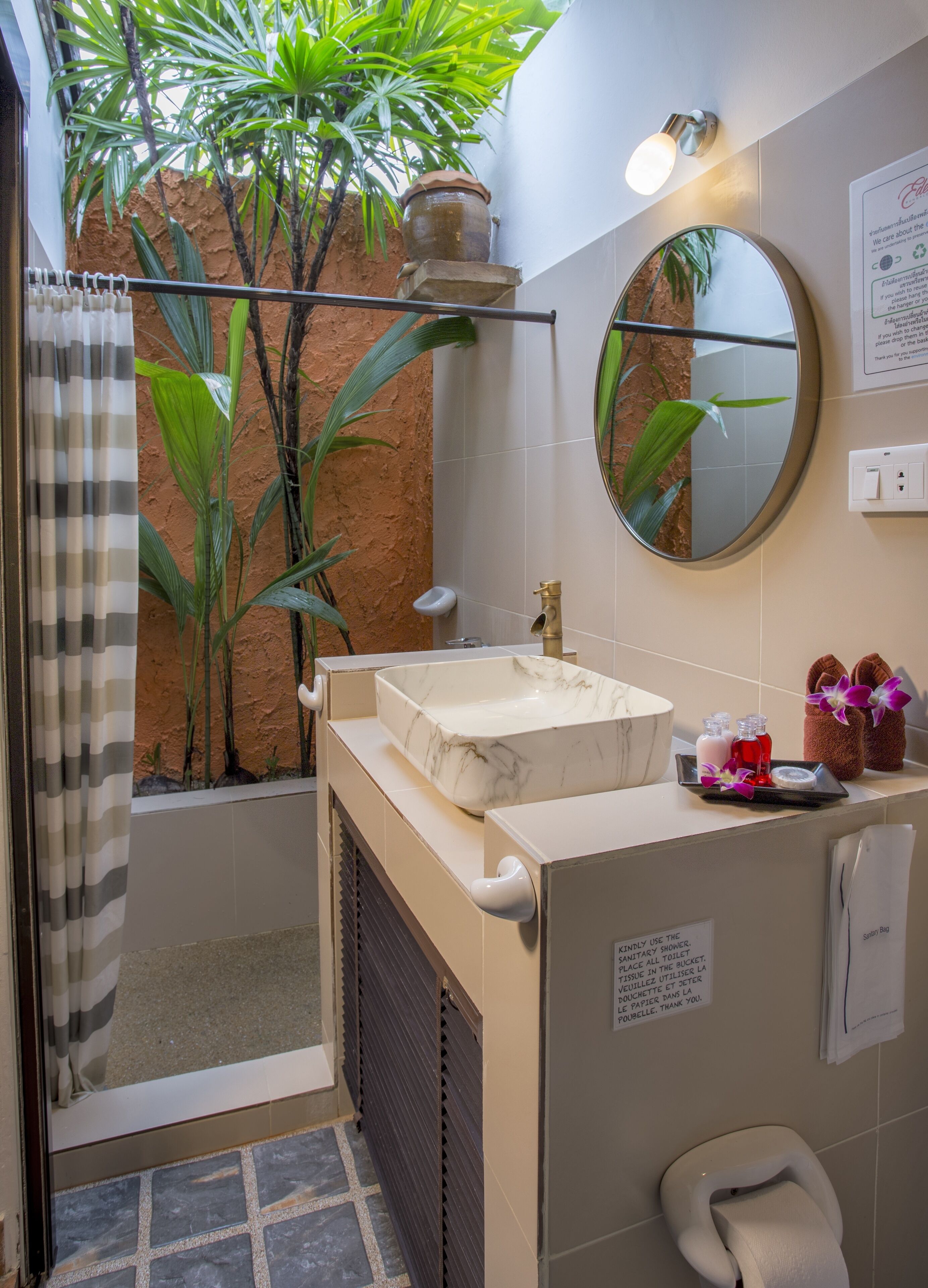 A bathroom with a shower, sink, and toilet. There are plants, a mirror, and toiletries on the counter.