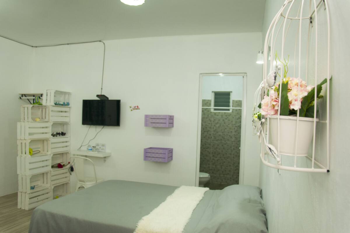 A bedroom with a bed, a TV mounted on the wall, a small shelf, and a white crib with storage drawers.