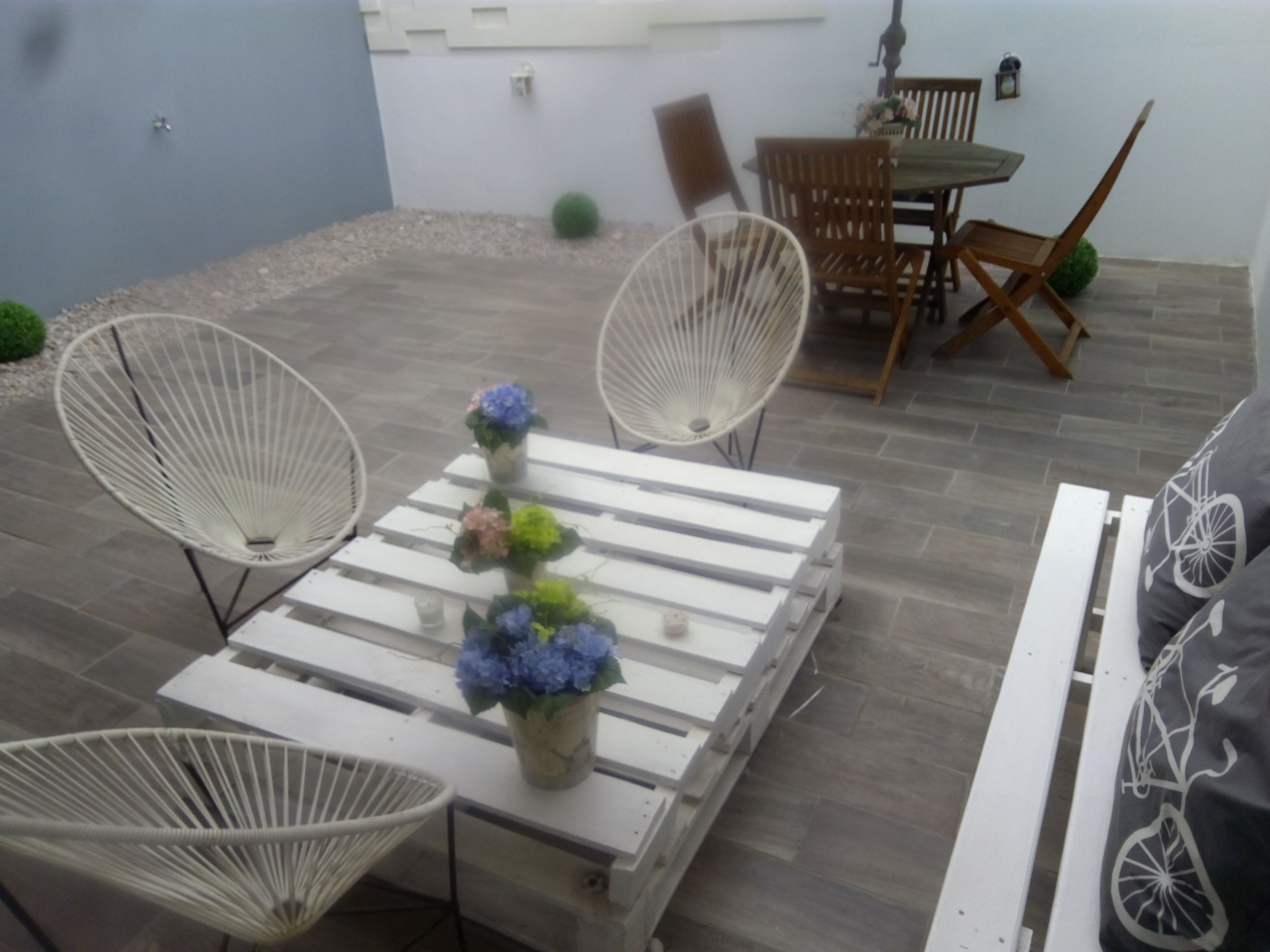 Um pátio com cadeiras de vime branco, uma mesa branca com um vaso de flores e uma área de refeições com cadeiras e mesa de madeira.
