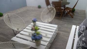 Um pátio com cadeiras de vime branco, uma mesa branca com um vaso de flores e uma área de refeições com cadeiras e mesa de madeira.