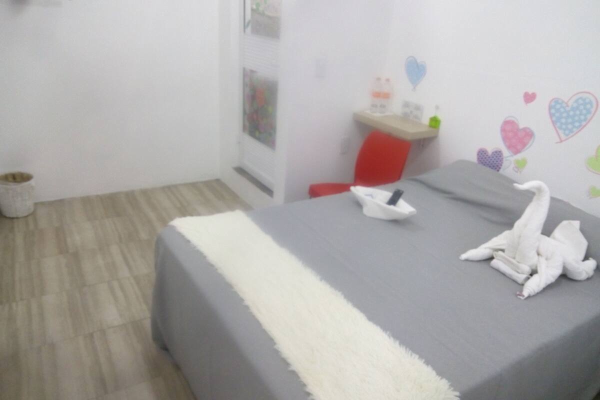 A neatly made bed with a grey bedspread and a white throw blanket, a red chair, a small desk with a bottle and a phone, and a wall decorated with heart stickers.