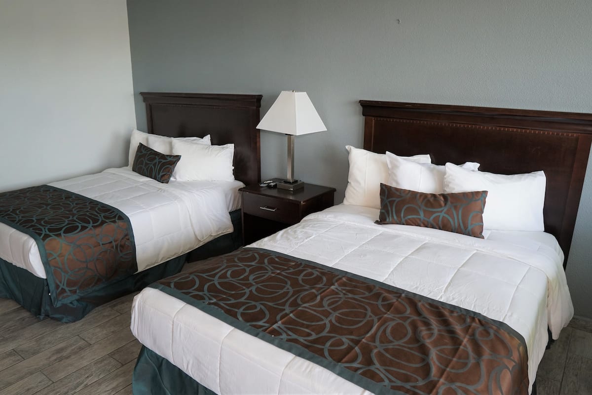 Habitación de hotel con dos camas, cada una con cabecero de madera, ropa de cama blanca y un plaid estampado. Hay una mesita de noche con una lámpara entre las camas.