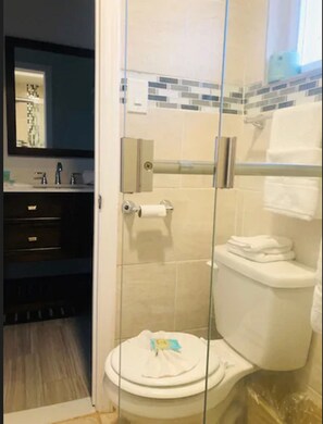 A bathroom with a glass-enclosed shower, a toilet with a paper towel holder, and a sink with a mirror above it. - Sunset Beach Hotel (Wildwood)