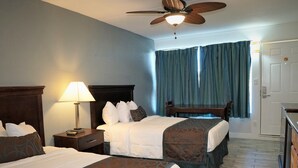 A hotel room with two beds, a desk, a ceiling fan, and a view of the outside through a window with curtains.