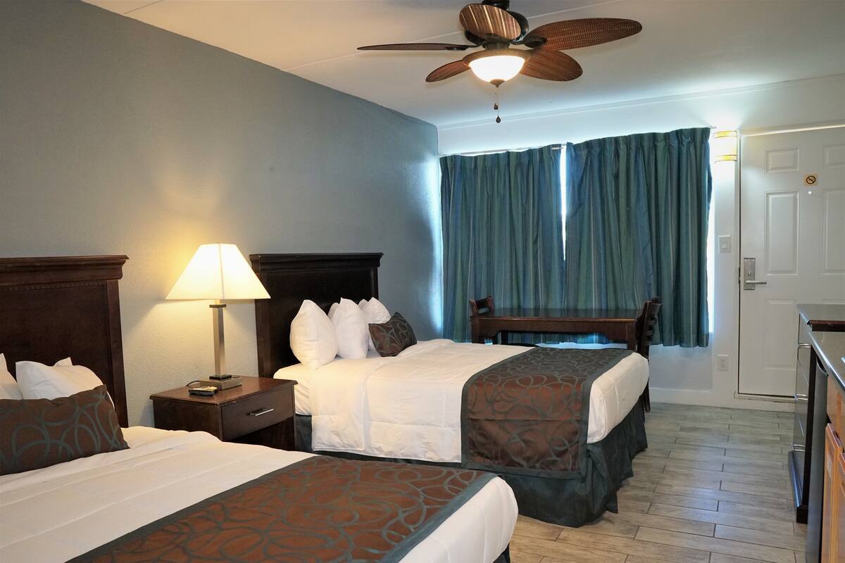 Habitación de hotel con dos camas, un escritorio, un ventilador de techo y vistas al exterior a través de una ventana con cortinas.