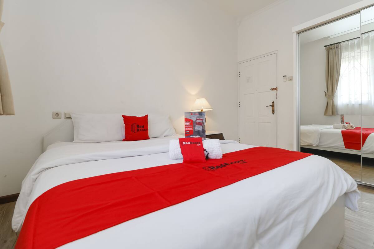 A hotel room with a bed, a red and white bedspread, a red door sign, a lamp, and a mirror.