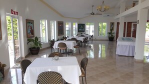 Uma ampla sala de jantar com várias mesas e cadeiras, grandes janelas e ventiladores de teto.