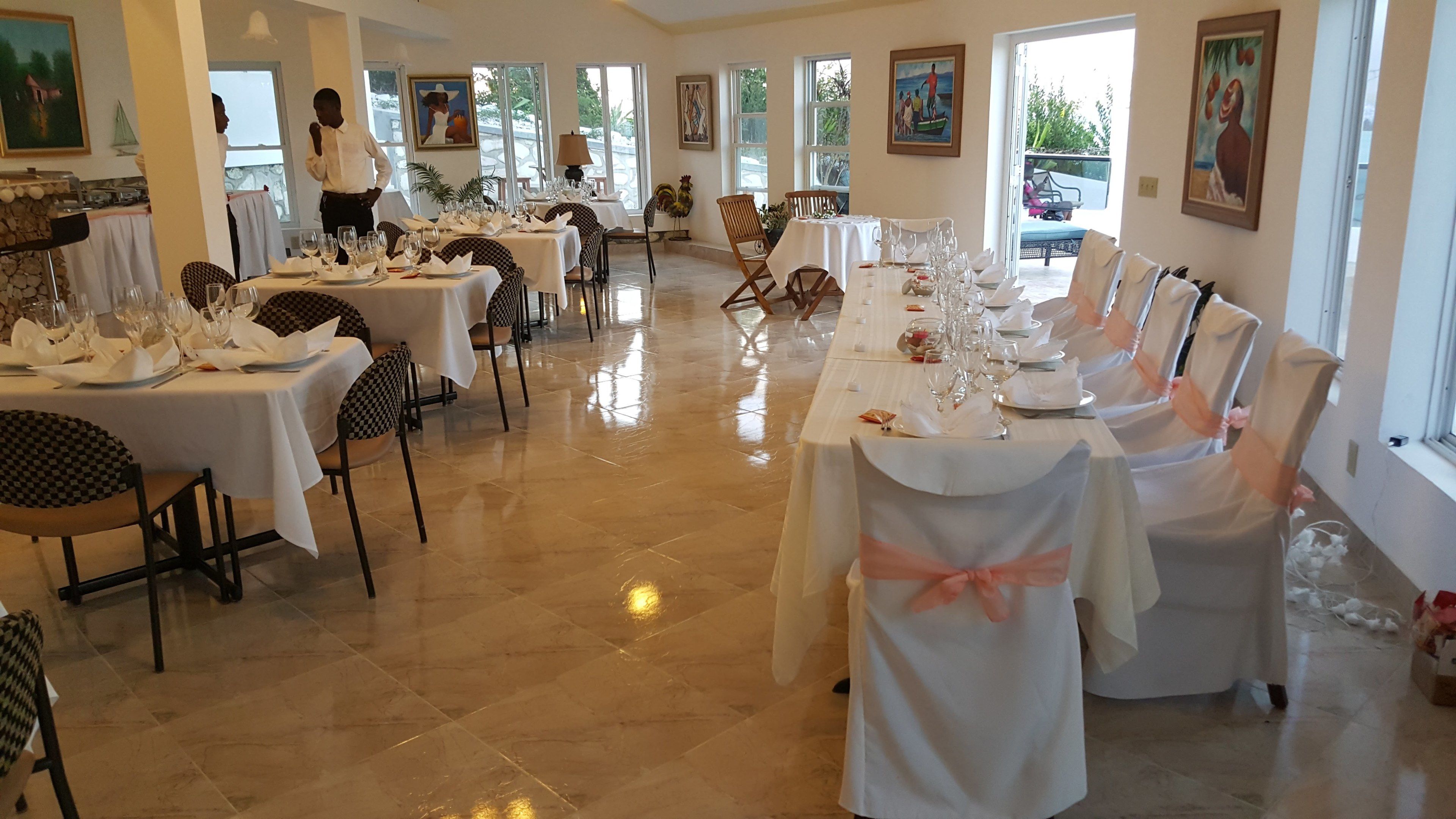 A spacious dining area with multiple tables set for a formal event, large windows allowing natural light, and artwork on the walls.