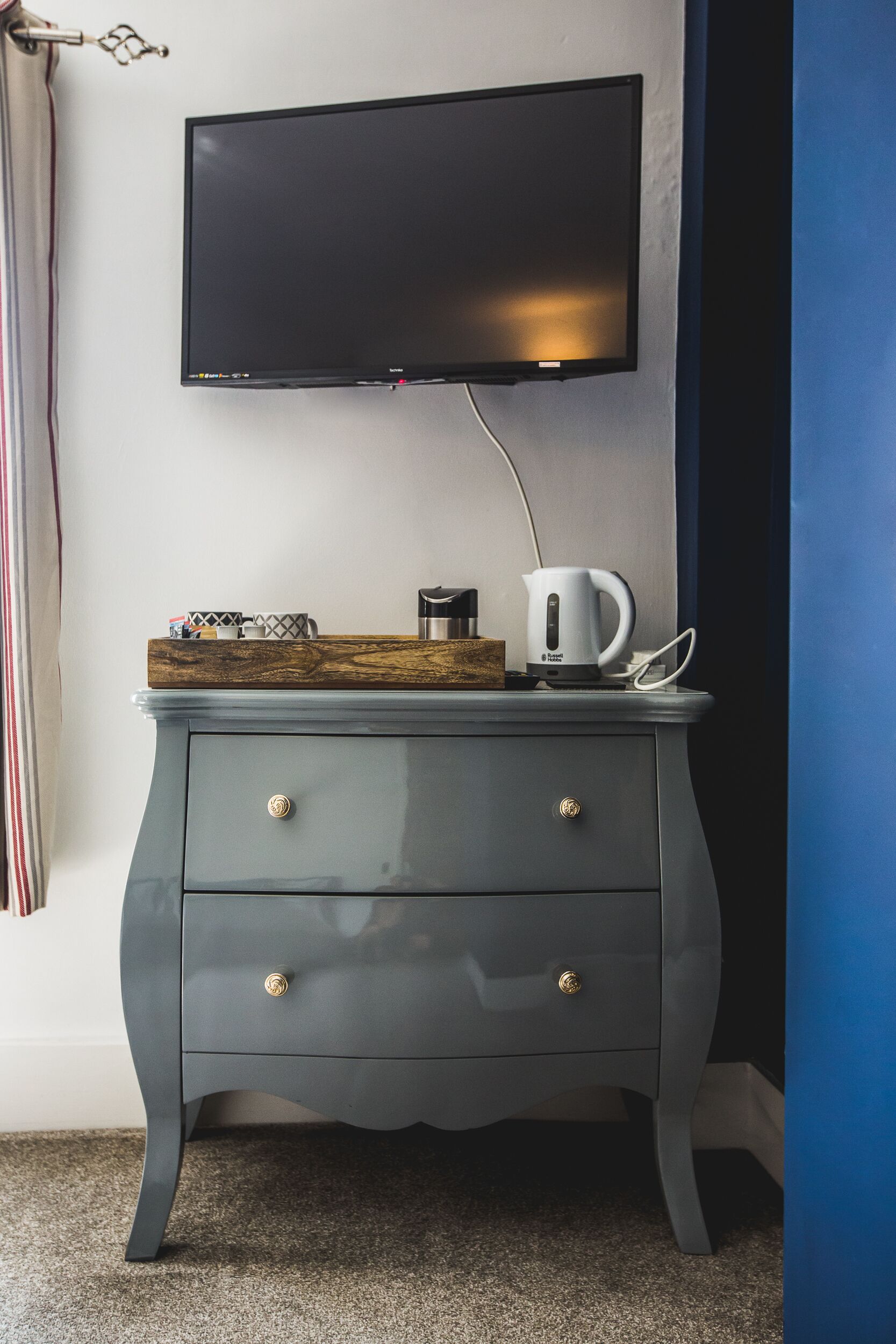 A modern room with a flat-screen TV mounted on the wall, a wooden tray with a coffee maker and mug, and a grey dresser with gold knobs.