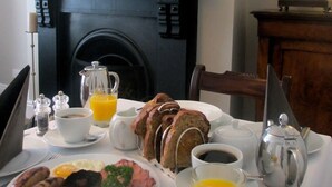 Une table de petit-déjeuner avec des assiettes remplies de nourriture, une cafetière, une théière et un pichet de jus d’orange.