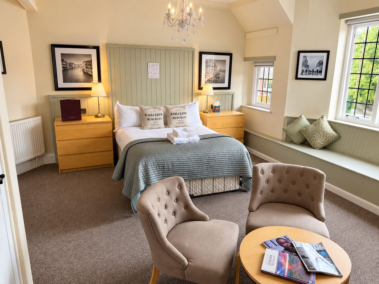 Een slaapkamer met een bed, twee nachtkastjes, een lampje, een raam met uitzicht, een kroonluchter en een zithoek met twee stoelen en een tafel.