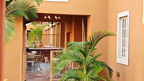 A covered outdoor seating area with a table and chairs, surrounded by palm trees and a building with a beige wall and a white window.