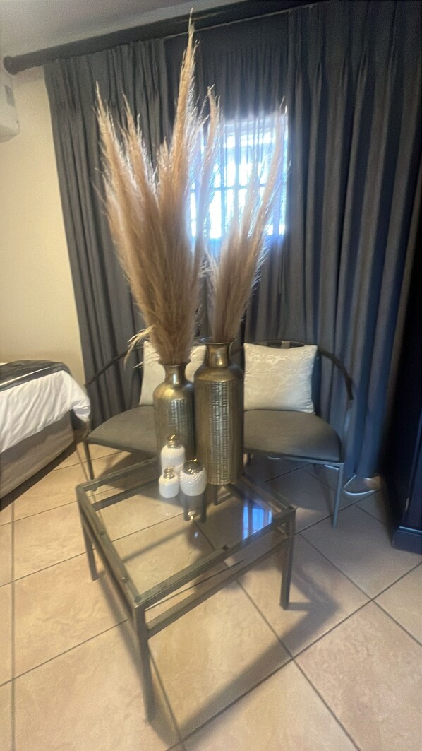 A room with a glass-top table, two vases with dried plants, a sofa, and a bed with a white cover.