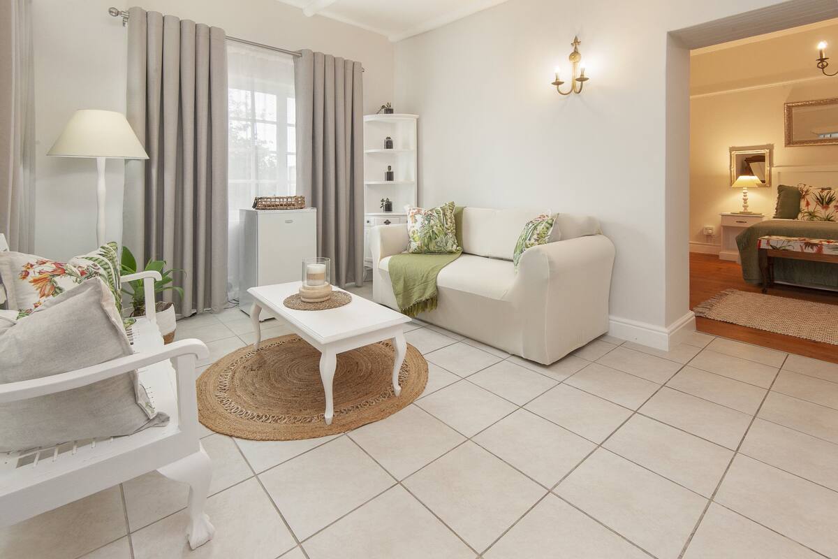 Sala de estar com um sofá branco, uma poltrona branca, uma mesinha com uma vela e um abajur. Há uma janela com cortinas, um armário branco e uma luminária na parede.