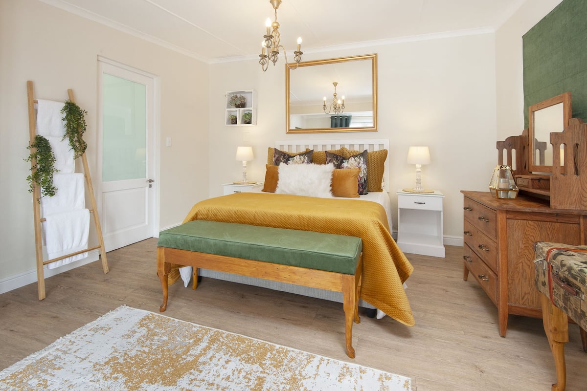Quarto com cama, um banquinho, cômoda, espelho e porta.