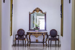 Una habitación con un espejo grande y ornamentado, dos sillones tapizados oscuros y una mesita con un jarrón de flores.