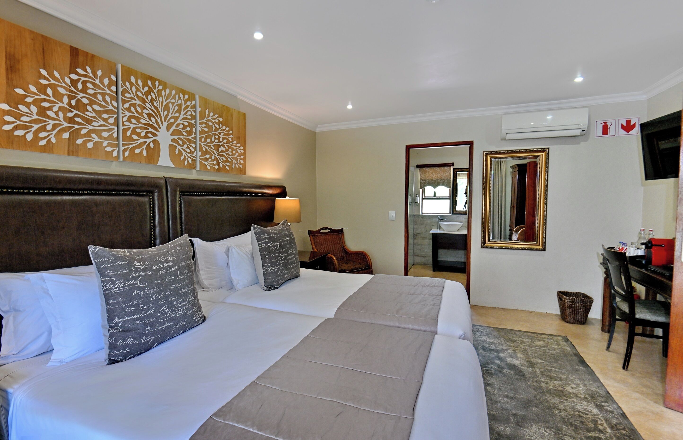 A hotel room with a large bed, a wooden headboard, a wall-mounted air conditioner, a mirror, and a desk with a chair.