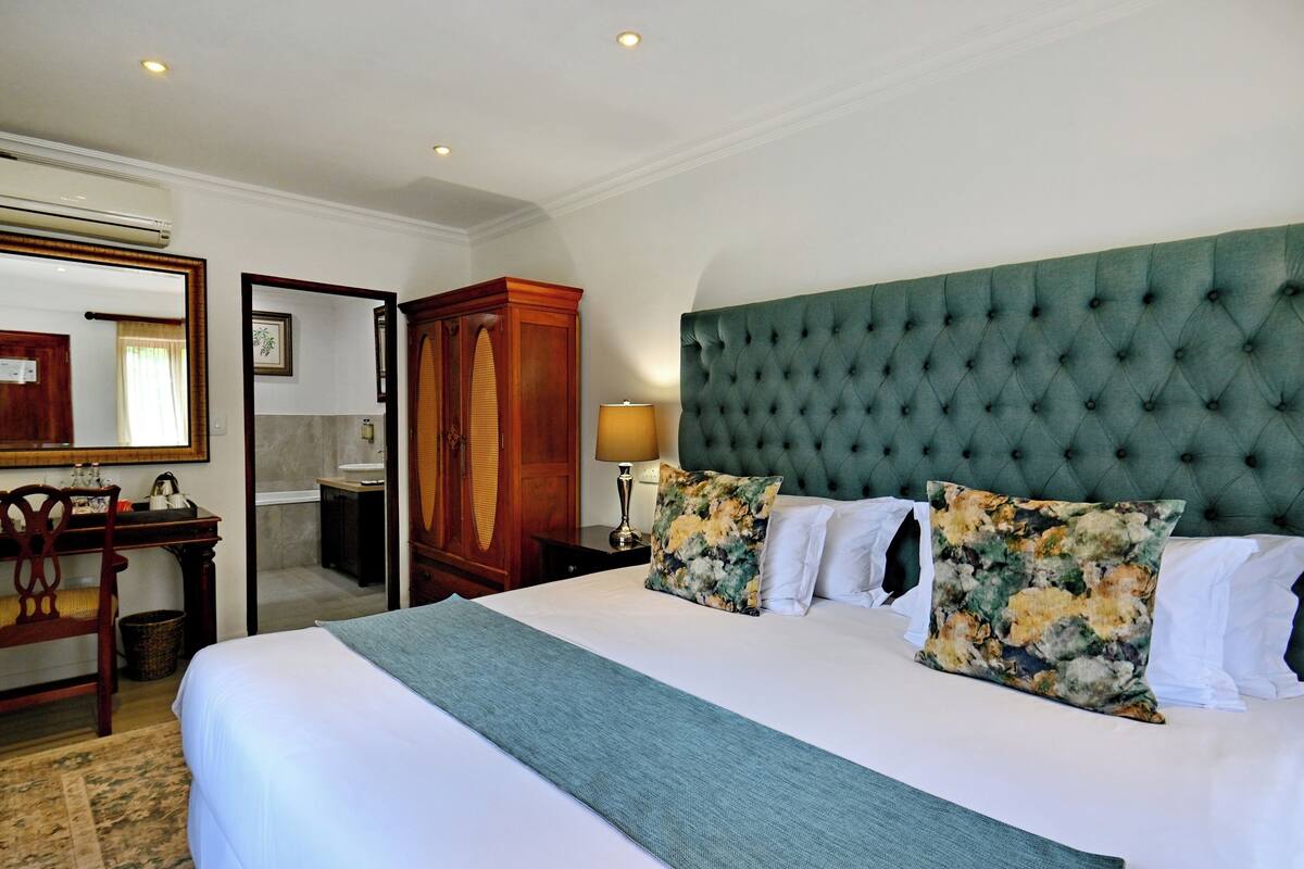 Une chambre d’hôtel avec un grand lit, une tête de lit recouverte d’un tissu vert, un bureau en bois et un miroir.
