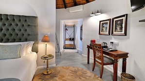 Quarto de hotel com cabeceira estofada, luminária de cabeceira, mesa de jantar com cadeiras e televisão na parede.