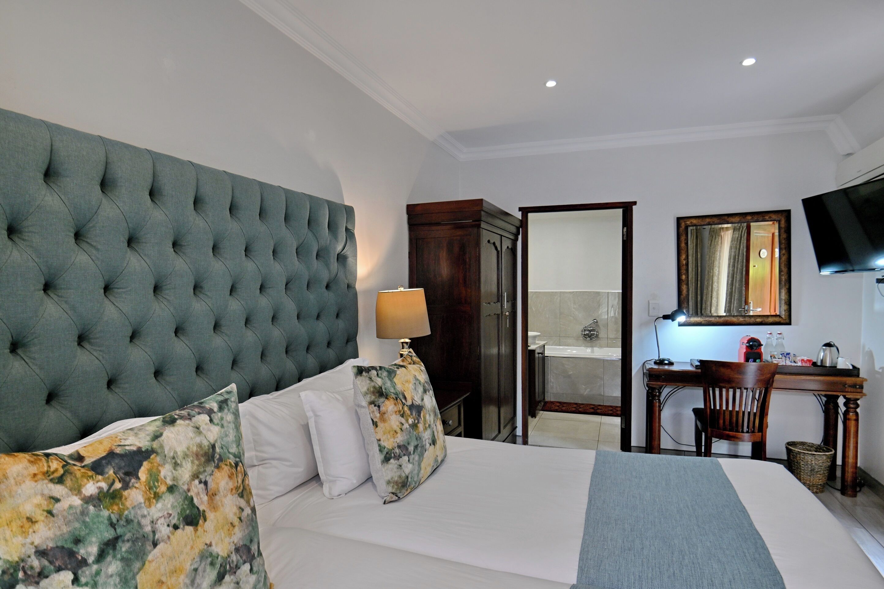 Quarto de hotel com uma grande cabeceira estofada, cama com lençóis brancos e almofadas florais, escrivaninha com luminária, televisão e espelho.