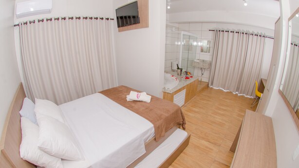 A modern hotel room with a large bed, a small desk, a mirror, and a shower area. - Pousada Rosa (Penha)