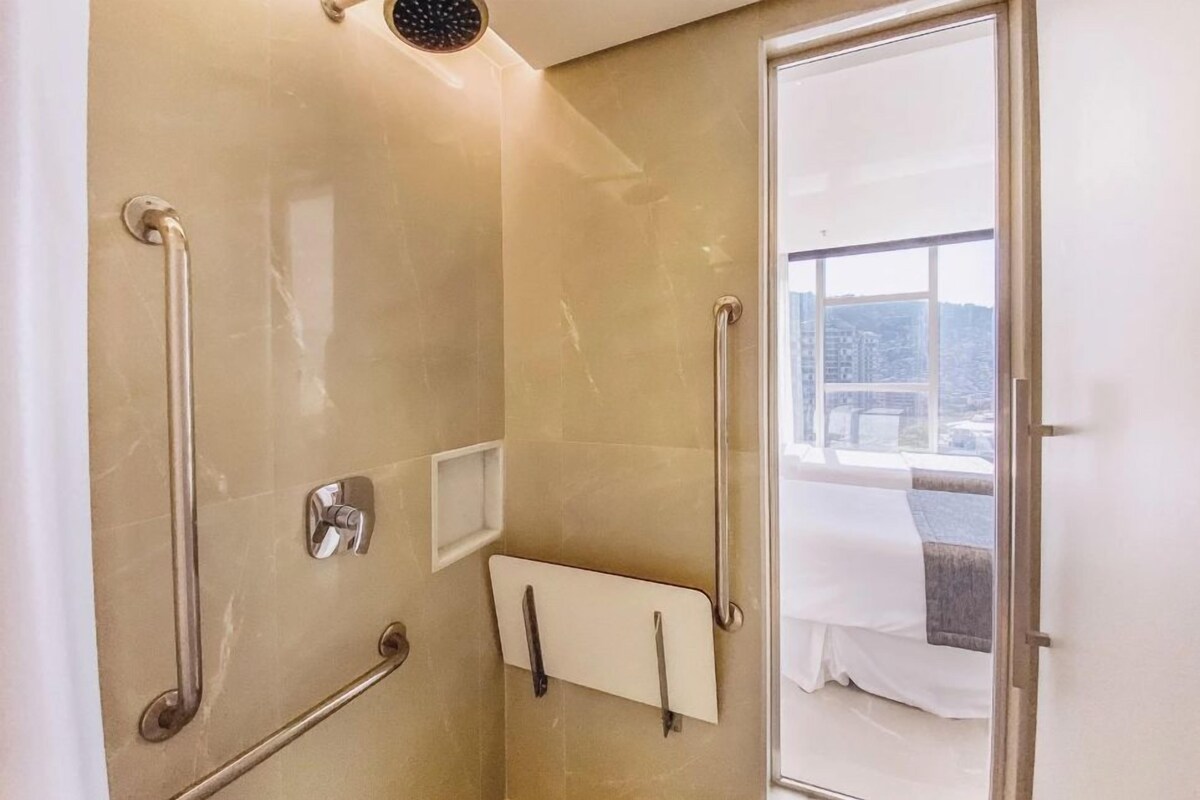 A bathroom with a shower, handrails, a towel rack, and a view of a cityscape through a glass door.