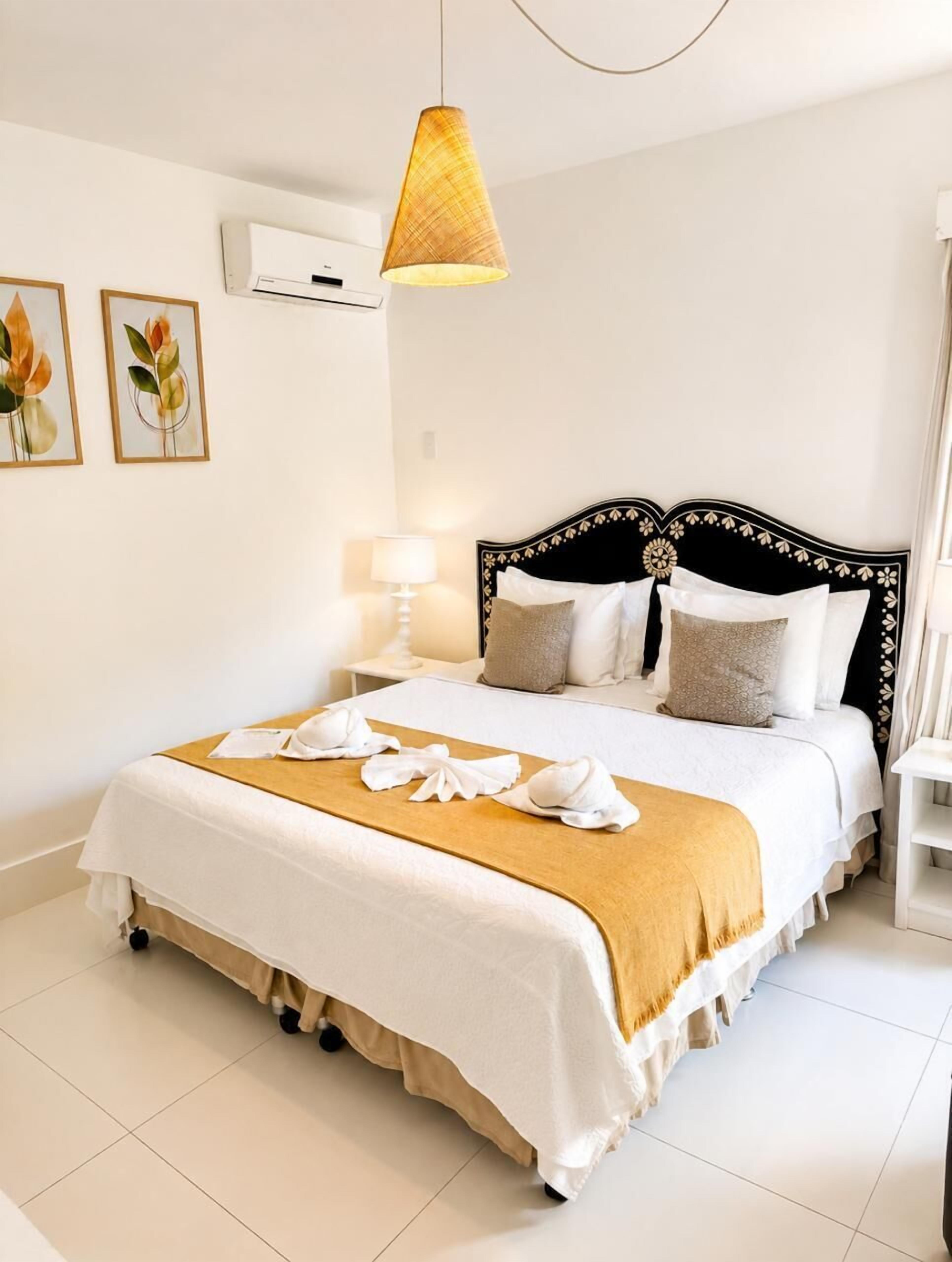 A neatly made bed with a white and beige bedspread, a gold throw blanket, and white towels arranged on the bed.